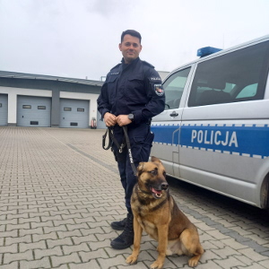 przewodnik psa służbowego wraz z psem na tle radiowozu.